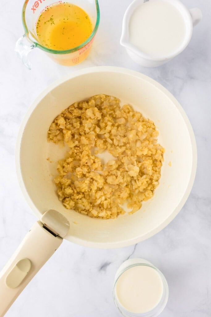 A white pan with sautéed onions, perfect for starting a green bean casserole, sits on a marble surface, surrounded by a measuring cup of broth and two small pitchers of milk or cream.