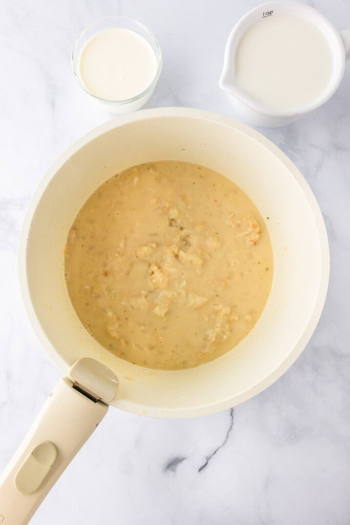 A white pan with a creamy sauce and chunks, perfect for green bean casserole, sits next to a small glass of cream and a white measuring cup filled with milk on a marble surface.