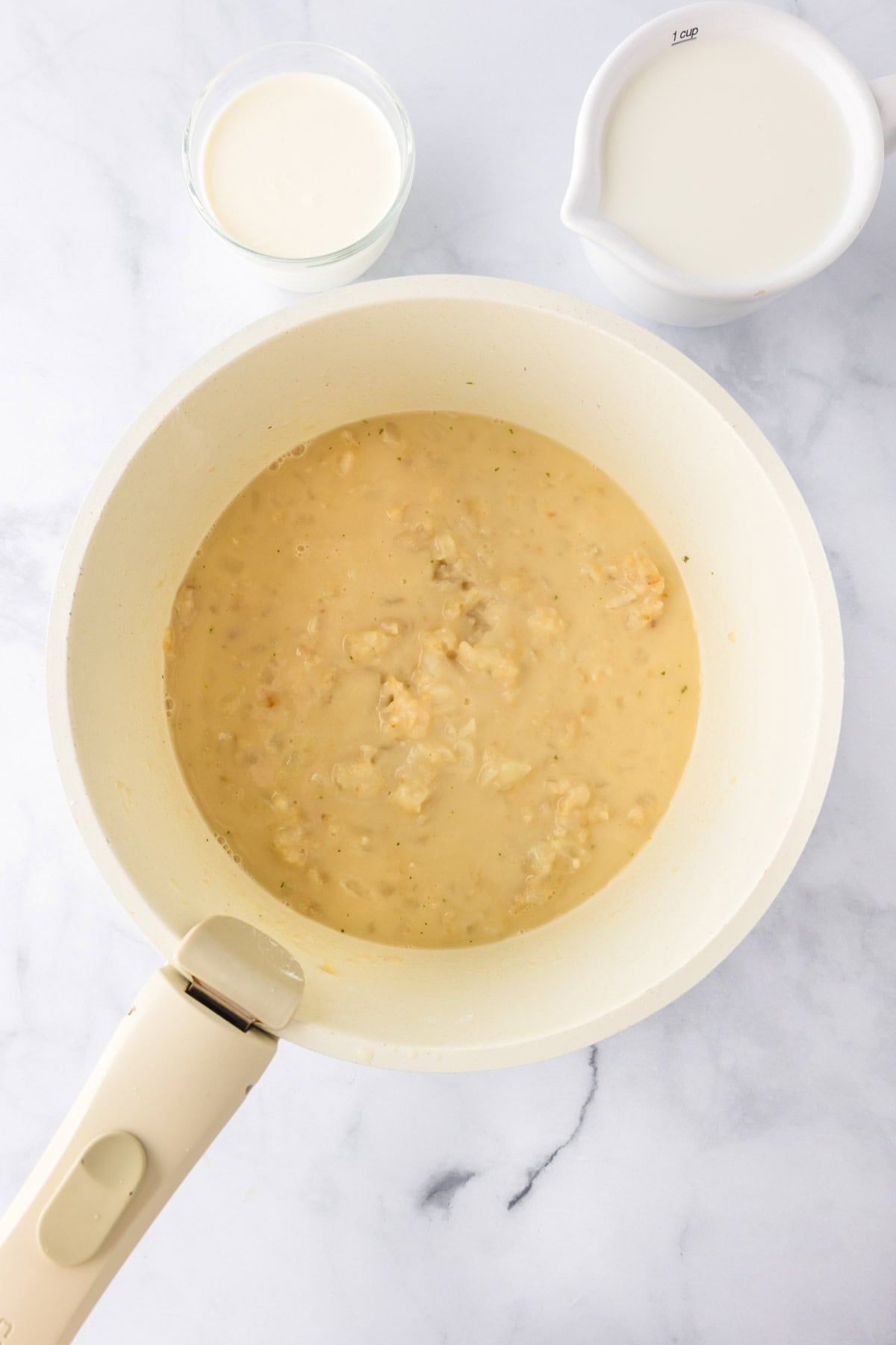 A white pan with a creamy sauce and chunks, perfect for green bean casserole, sits next to a small glass of cream and a white measuring cup filled with milk on a marble surface.