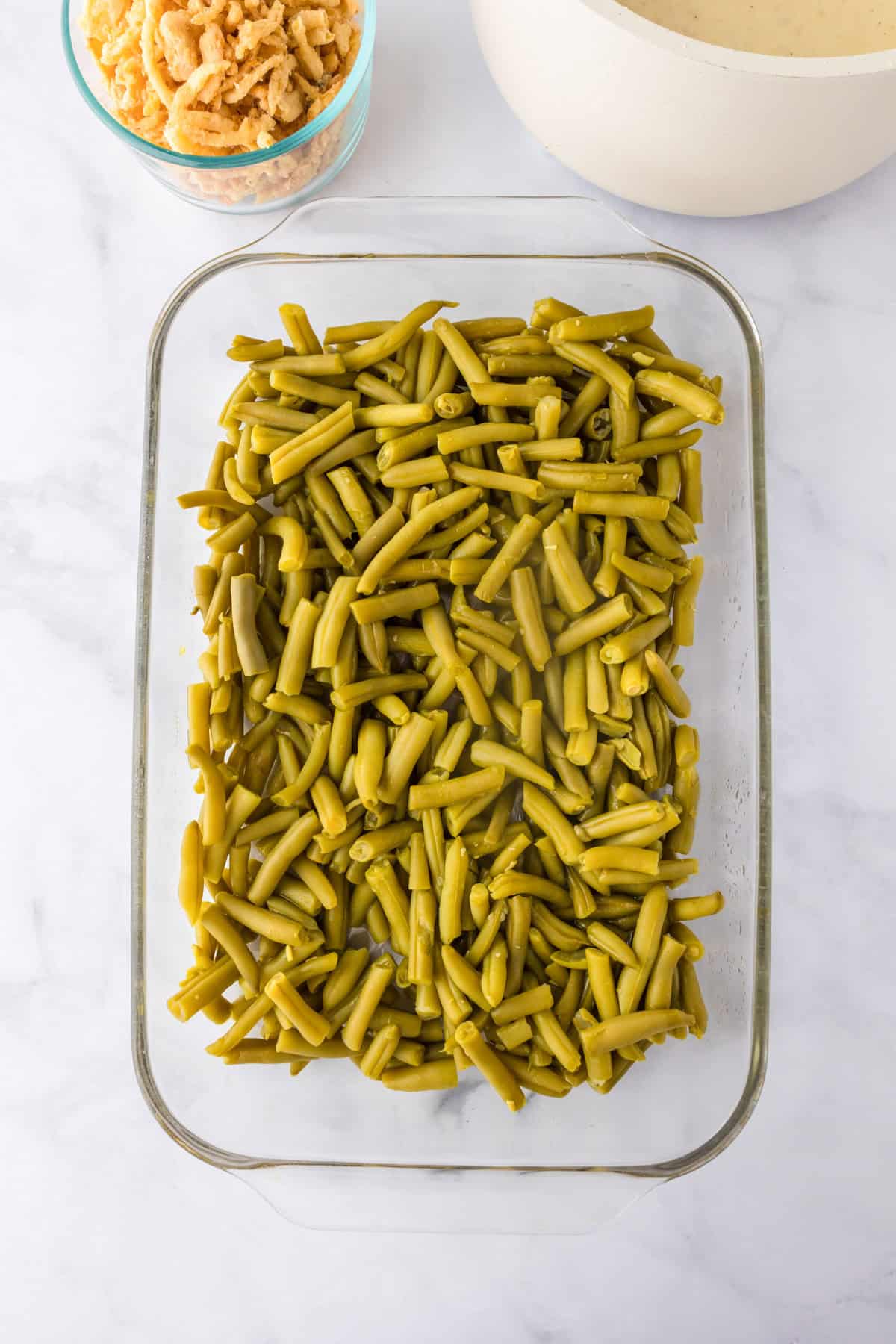 A glass baking dish filled with cut green beans on a marble surface, alongside a bowl of crispy fried onions and sauce—everything you need to make the perfect green bean casserole.