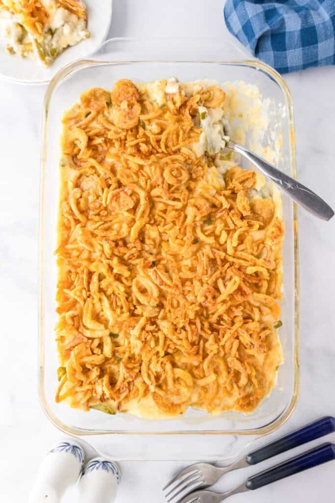 A glass baking dish filled with green bean casserole topped with crispy fried onions, a serving spoon inside. A plated serving of the classic green bean casserole and utensils are nearby.