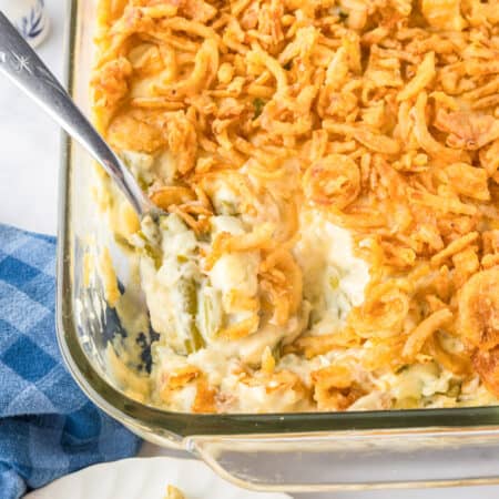 A glass baking dish of green bean casserole topped with crispy fried onions, with a serving spoon partially scooping out the savory casserole.