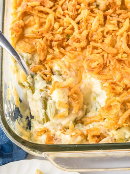A glass baking dish of green bean casserole topped with crispy fried onions, with a serving spoon partially scooping out the savory casserole.