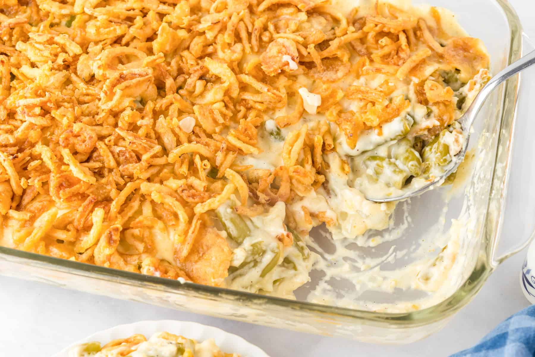 A glass baking dish of green bean casserole topped with crispy fried onions, partially served with a spoon in the dish. This classic green bean casserole looks delicious and ready to enjoy.
