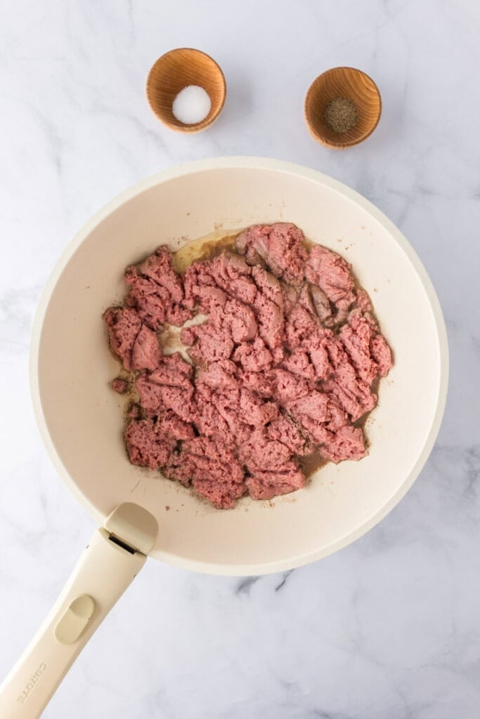 Ground turkey spaghetti is being prepared as ground meat sizzles in a white skillet on a marble surface, with small wooden bowls of salt and pepper nearby.