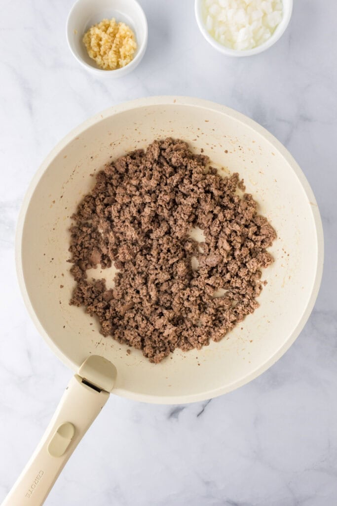 A white skillet with browned ground turkey for spaghetti on a marble surface, accompanied by small bowls of chopped onions and minced garlic in the background.