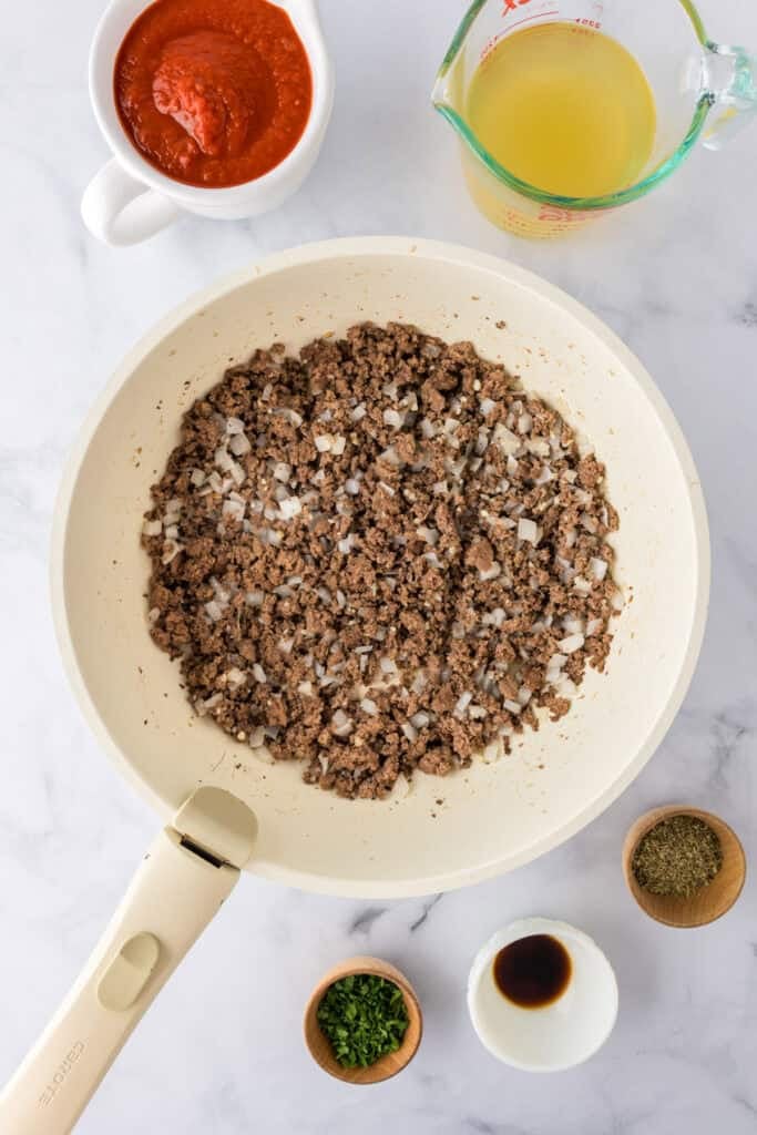 A pan with browned ground turkey and onions sits on a counter surrounded by tomato sauce, broth, dried herbs, Worcestershire sauce, and chopped parsley for making ground turkey spaghetti.