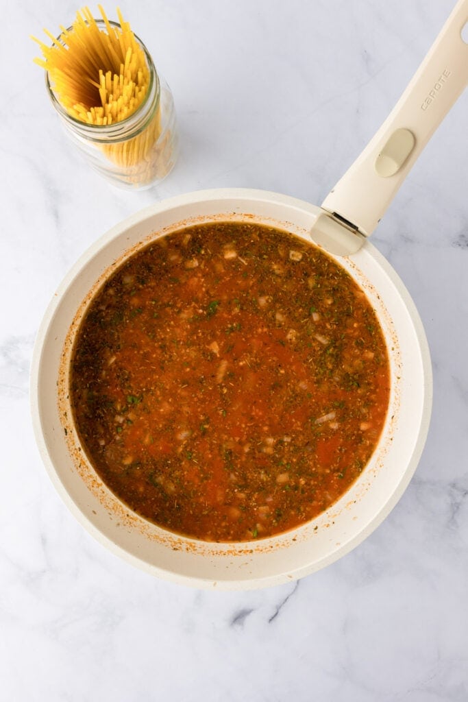 A white pan filled with ground turkey and sauce sits on a marble surface next to a jar of uncooked spaghetti noodles for ground turkey spaghetti.