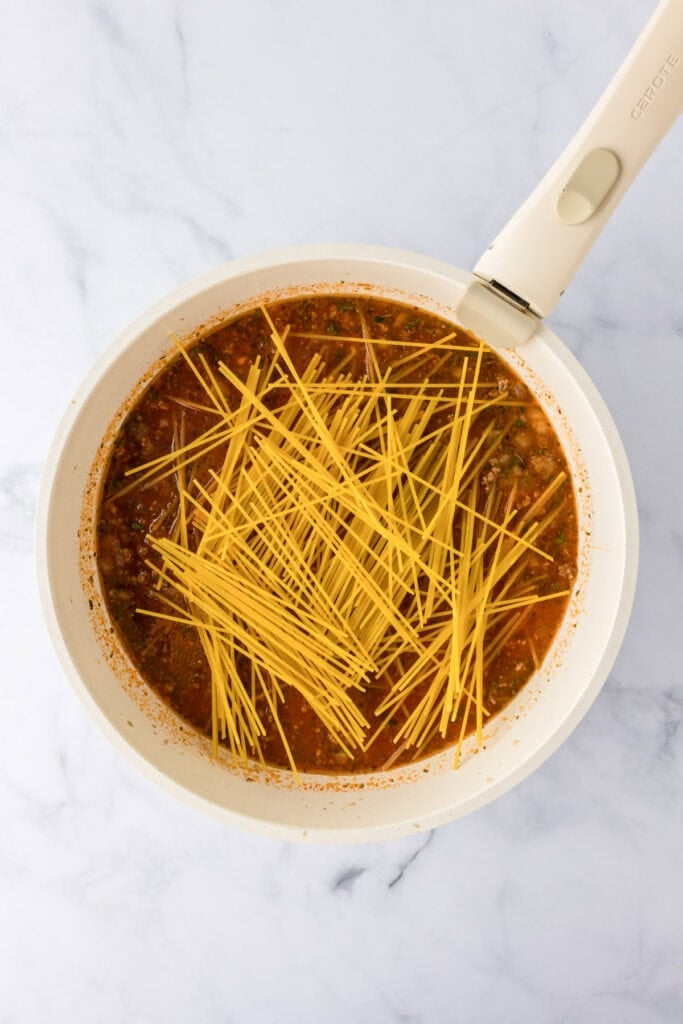 Uncooked spaghetti noodles in a white pan with tomato sauce and ground turkey, viewed from above, making a delicious ground turkey spaghetti.