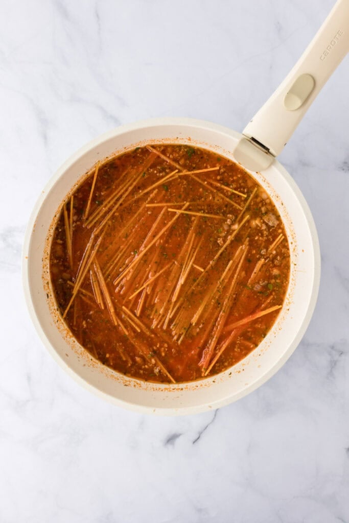 A white pan filled with uncooked ground turkey spaghetti in a red tomato-based sauce and broth, resting on a marble surface.