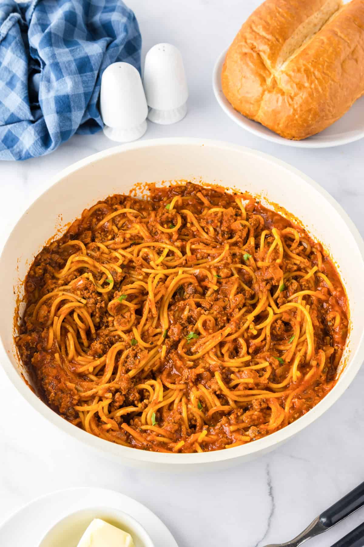 A skillet of ground turkey spaghetti with meat sauce sits on a marble surface, accompanied by a loaf of bread, butter, salt and pepper shakers, and a blue checkered cloth nearby.