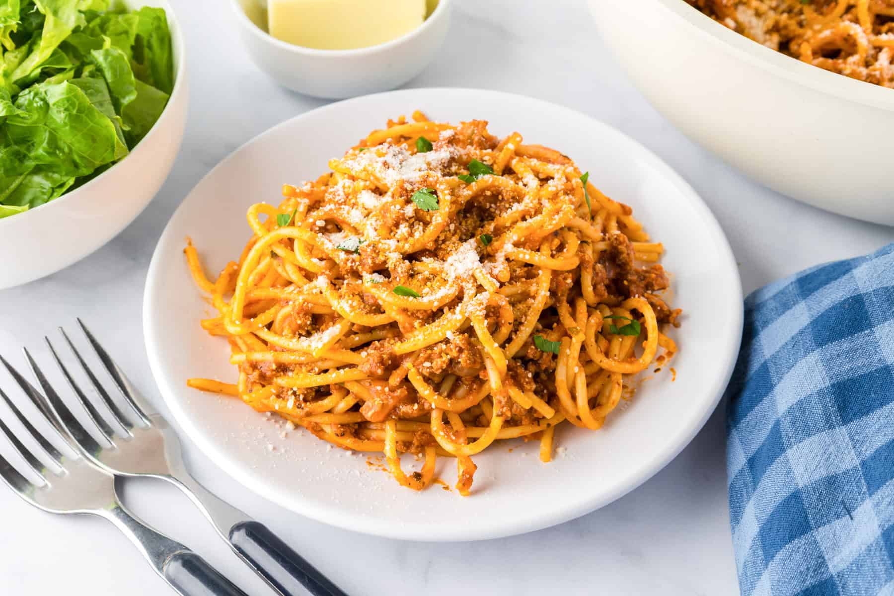 A white plate of ground turkey spaghetti with meat sauce and grated cheese, garnished with herbs, sits on a table next to forks, a bowl of lettuce, and a blue checkered napkin.