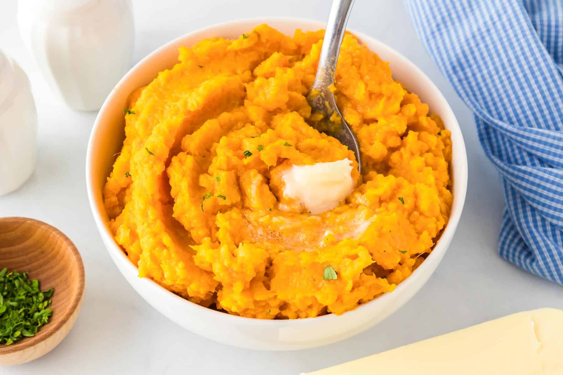 A bowl of creamy mashed sweet potatoes with a pat of butter melting on top, a spoon nestled in the bowl, and a blue striped cloth nearby.