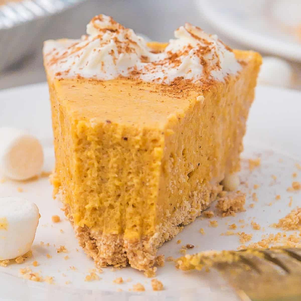 A slice of pumpkin marshmallow pie with whipped cream and cinnamon on top, sitting on a white plate surrounded by pie crumbs and mini marshmallows.