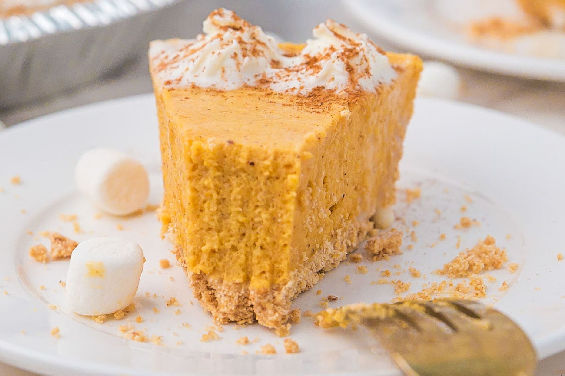 Slice of pumpkin marshmallow pie with whipped cream and a sprinkle of cinnamon on top, served on a white plate with mini marshmallows and crumbs.