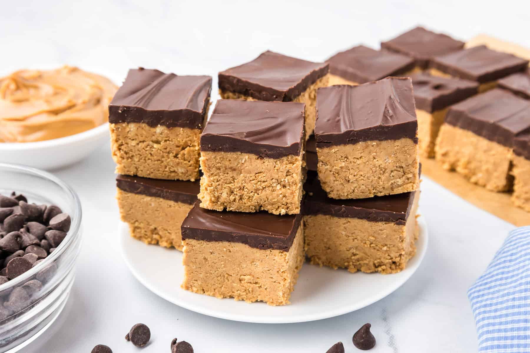 A plate of chocolate-topped peanut butter squares is stacked on a white surface, with bowls of chocolate chips and peanut butter nearby.
