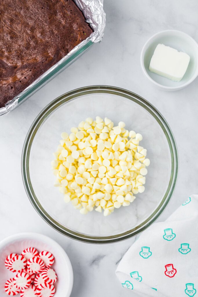 A glass bowl of white chocolate chips sits on a counter next to a pan of peppermint bark brownies, a small dish of butter, and a bowl of peppermint candies.
