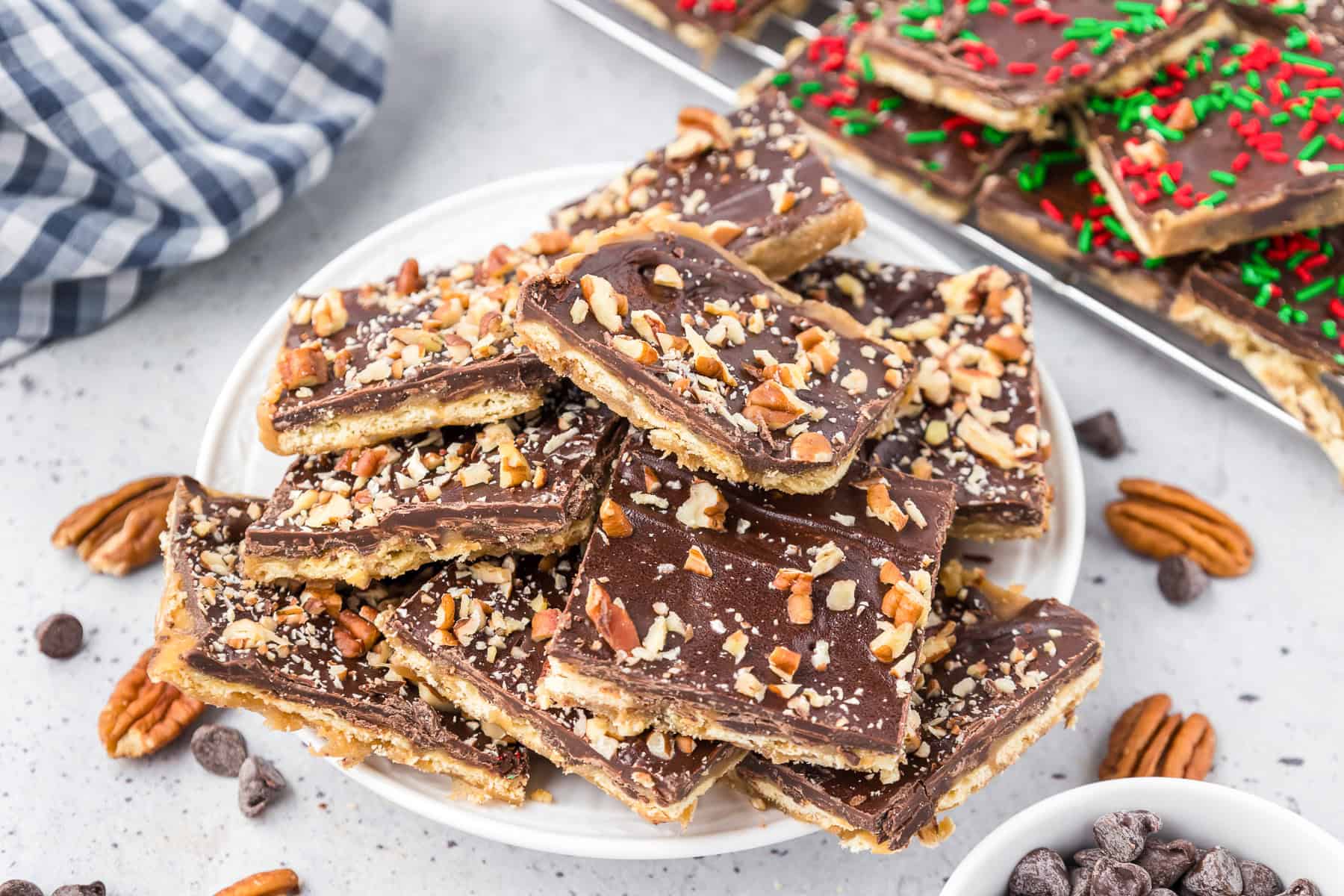A plate of chocolate toffee bars, made with crisp saltine cracker toffee and topped with chopped pecans, sits on a table surrounded by chocolate chips, pecan halves, and a tray of decorated bars in the background.