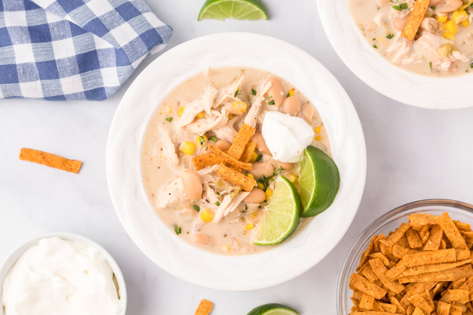 A bowl of creamy slow cooker white chicken chili topped with tortilla strips, a dollop of sour cream, and lime wedges, with extra sour cream and tortilla strips on the side.