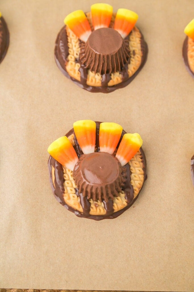 A festive turkey cookie decorated with a chocolate peanut butter cup, candy corn feathers, and melted chocolate on a round cookie, all arranged on parchment paper.