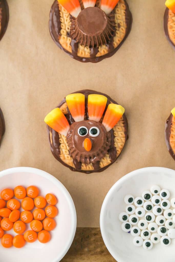 A turkey cookie decorated with candy corn feathers, candy eyes, and an orange chocolate beak is surrounded by bowls of orange candies and extra candy eyes.