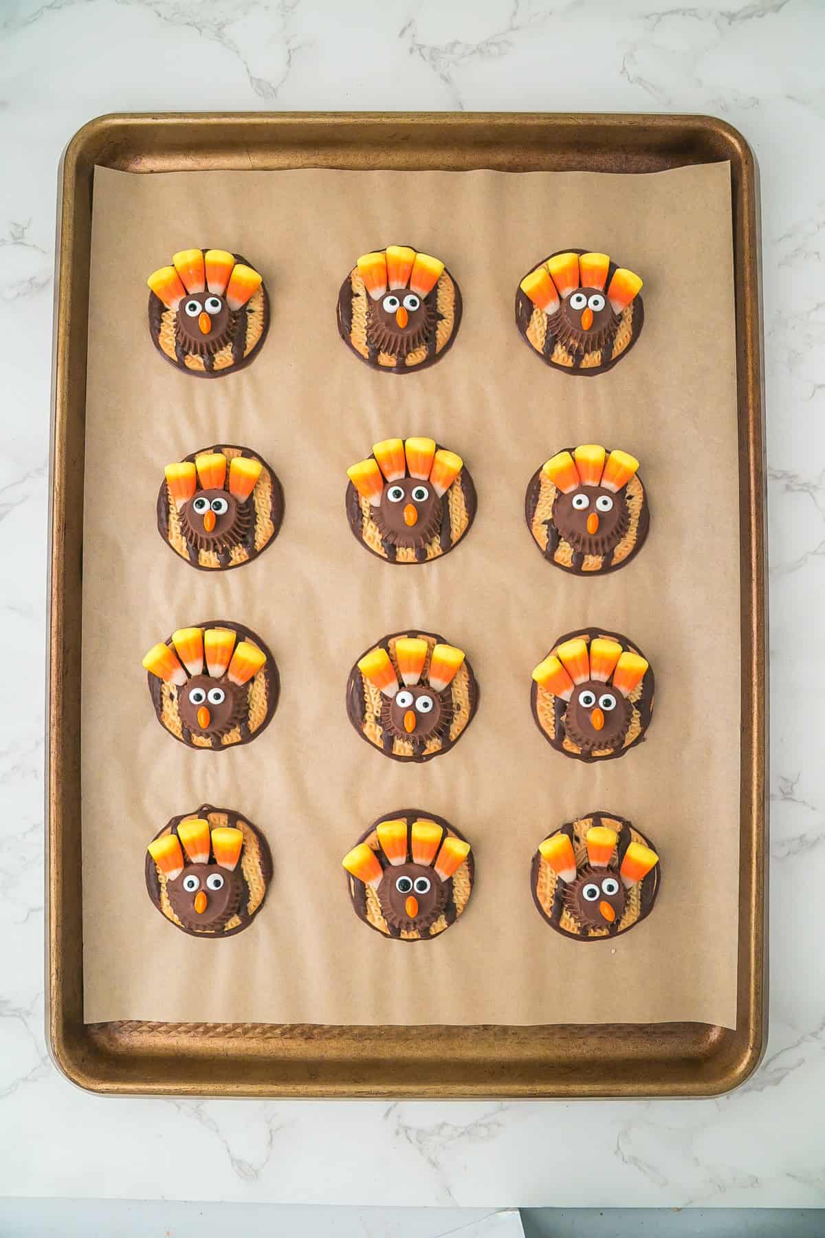 A baking tray with twelve turkey cookies decorated to look like turkeys using candy corn for feathers and small candy eyes, arranged in rows on parchment paper.