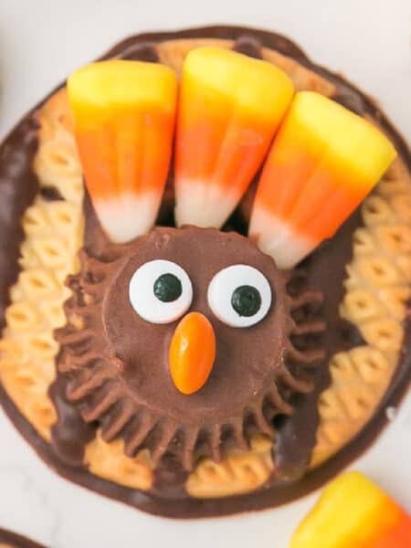 A turkey cookie decorated with a peanut butter cup, candy eyes, an orange candy beak, and three candy corn pieces as colorful feathers.