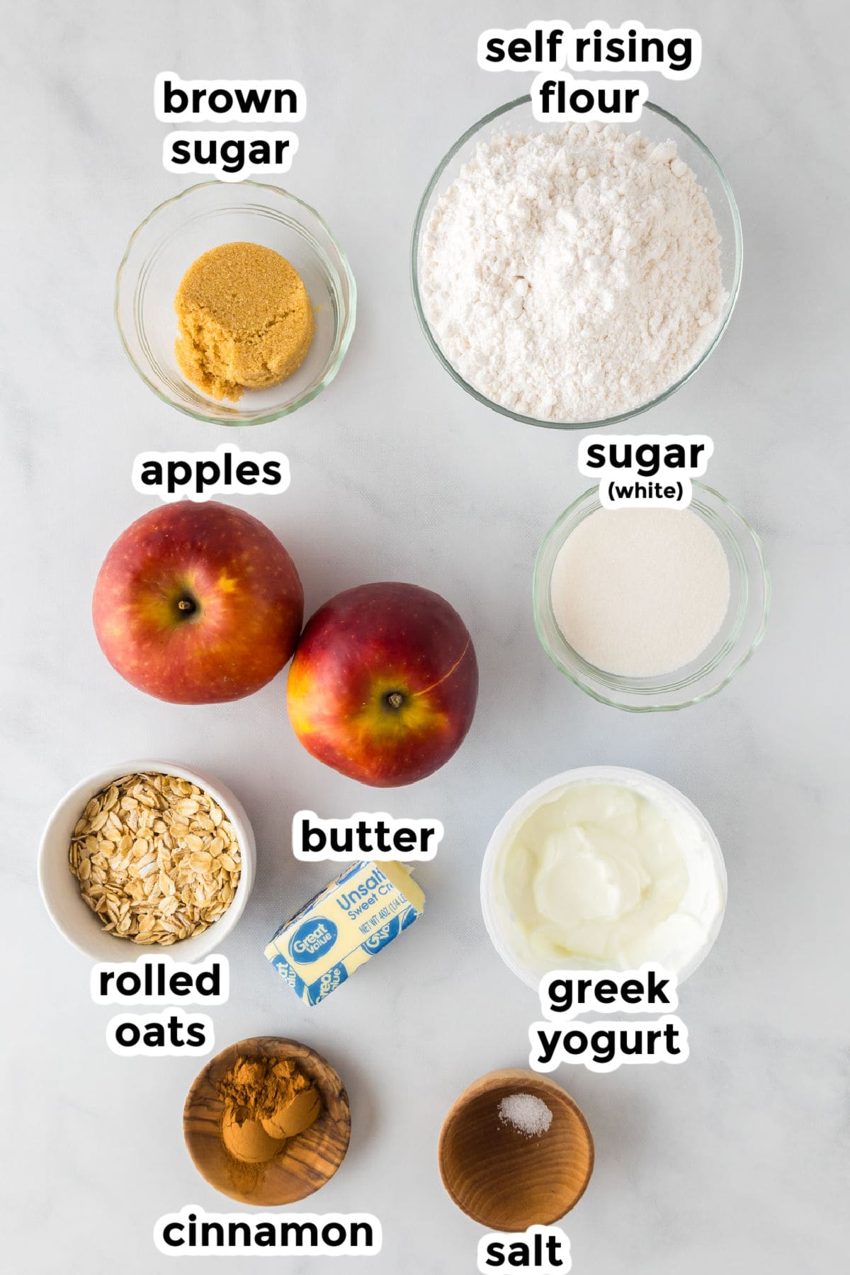 Top-down view of labeled baking ingredients for an apple dessert pizza, including brown sugar, self-rising flour, sugar, apples, rolled oats, butter, Greek yogurt, cinnamon, and salt on a white surface.