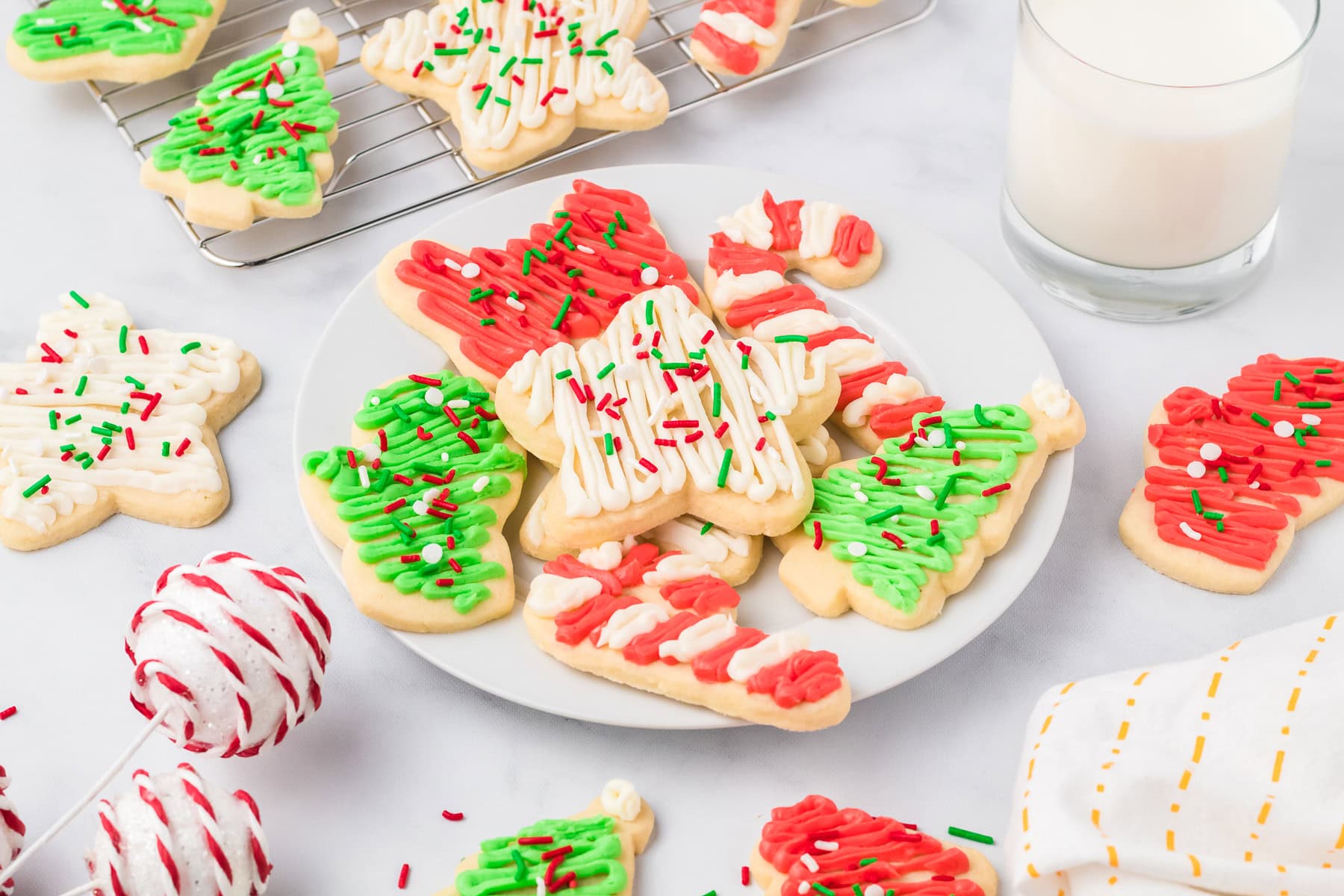 Plate of Christmas frosted sugar cookies with colorful icing and sprinkles sits next to a glass of milk, festive decorations, and a cooling rack filled with more cookies in the background.