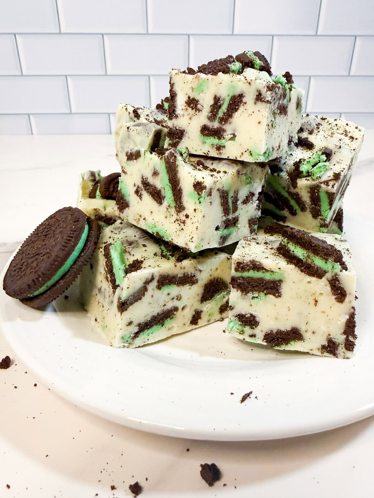 A plate stacked with pieces of creamy mint Oreo fudge, packed with crushed mint chocolate sandwich cookies, with whole and broken cookies scattered around.