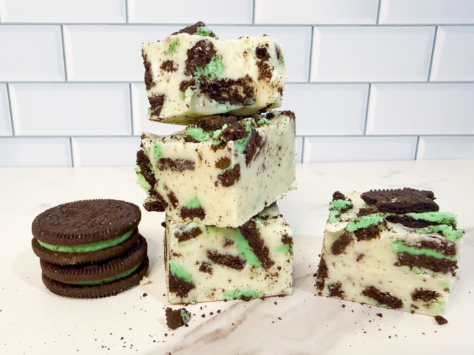 Four pieces of mint Oreo fudge with green and chocolate cookie chunks are stacked on a counter beside a stack of mint chocolate sandwich cookies, against a white tiled background.