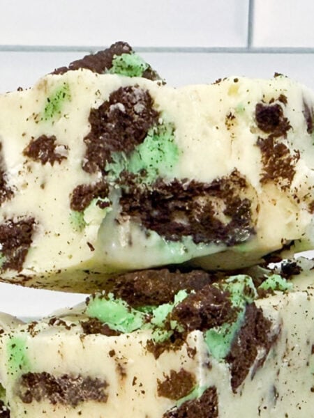 Two stacked pieces of mint Oreo fudge with chunks of green mint and chocolate cookies, set against a white tiled background.