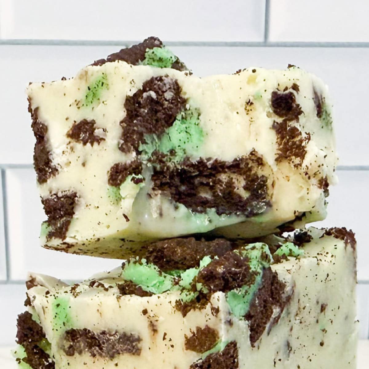Two stacked pieces of mint Oreo fudge with chunks of green mint and chocolate cookies, set against a white tiled background.