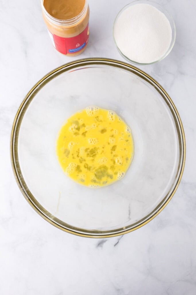 A glass bowl with beaten eggs sits on a white surface; nearby are a jar of peanut butter and a bowl of sugar—classic fixings for 3 ingredient peanut butter cookies.