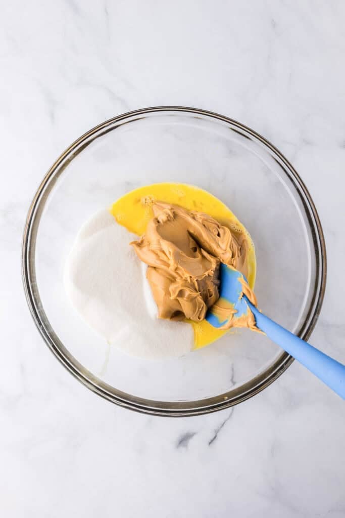 A glass bowl containing beaten eggs, granulated sugar, and creamy peanut butter for 3 ingredient peanut butter cookies is being mixed with a blue spatula on a white surface.