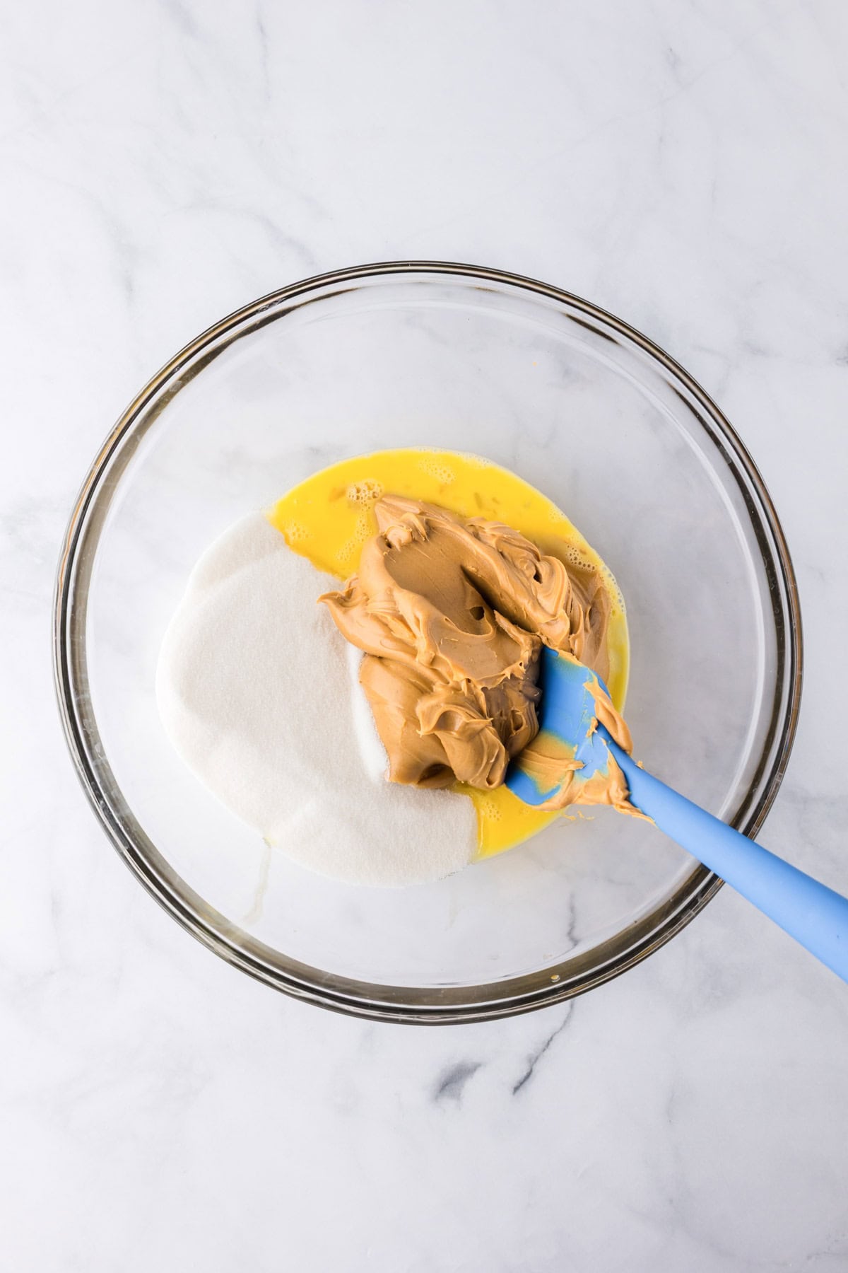 A glass bowl containing beaten eggs, granulated sugar, and creamy peanut butter for 3 ingredient peanut butter cookies is being mixed with a blue spatula on a white surface.