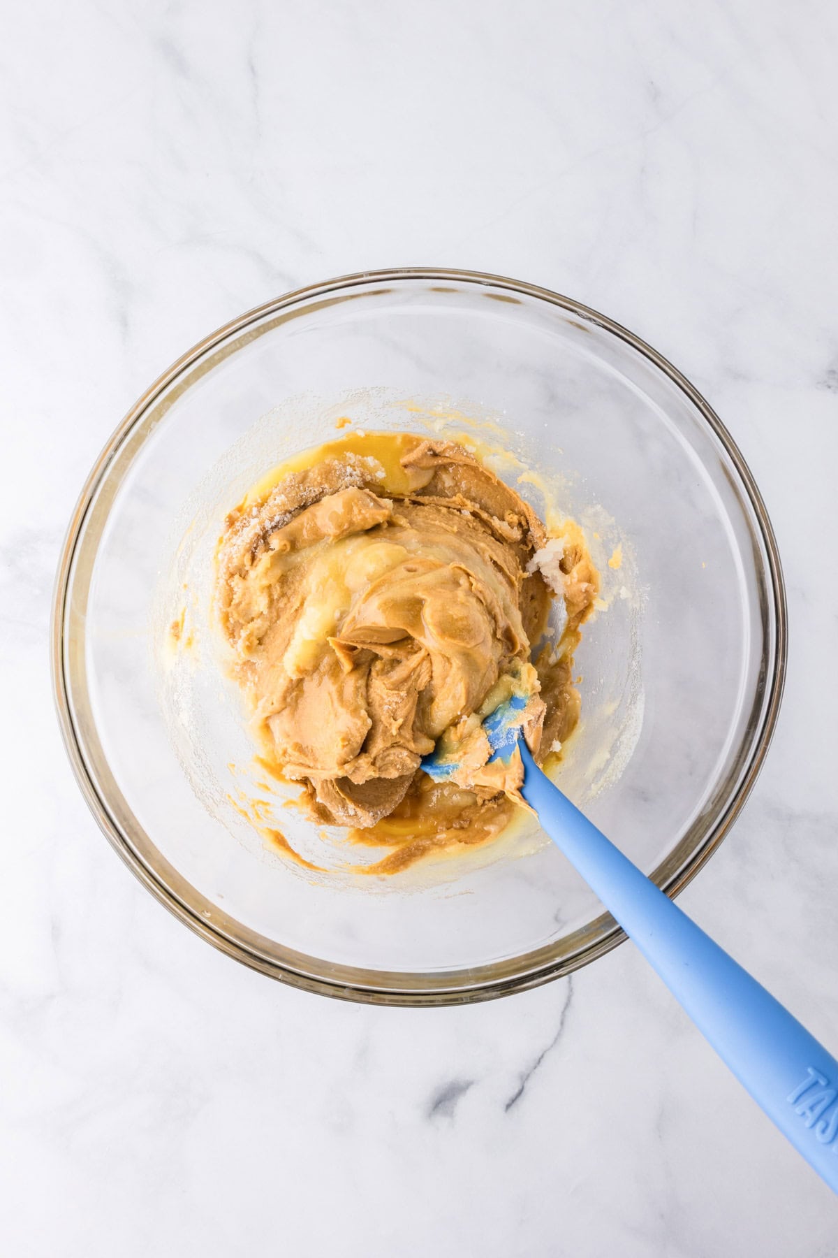 A glass bowl containing a thick, light brown 3 ingredient peanut butter cookie dough mixture being stirred with a blue spatula, placed on a white marble surface.