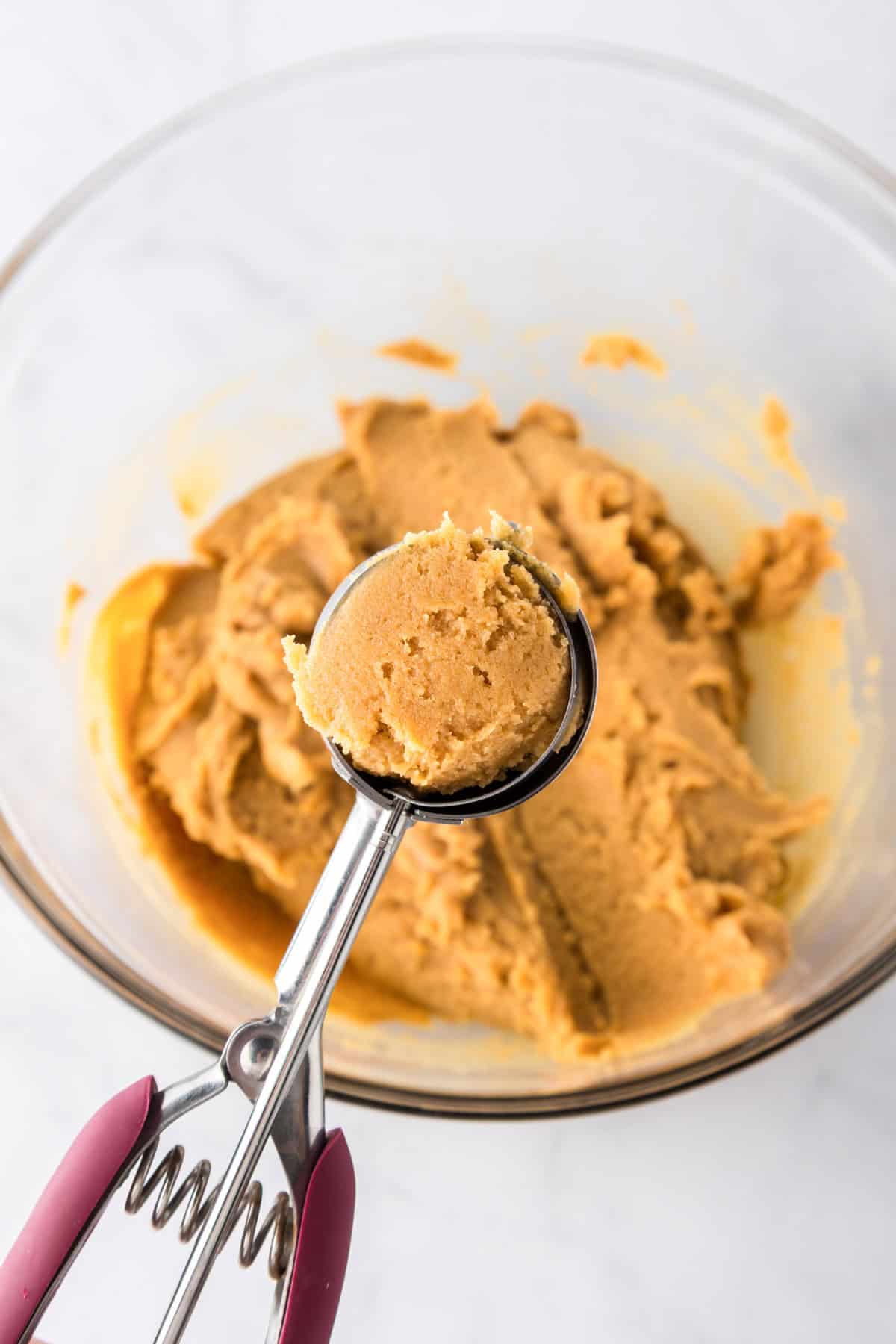 A cookie scoop holds a rounded portion of light brown 3 ingredient peanut butter cookies dough above a glass bowl containing more dough on a white surface.