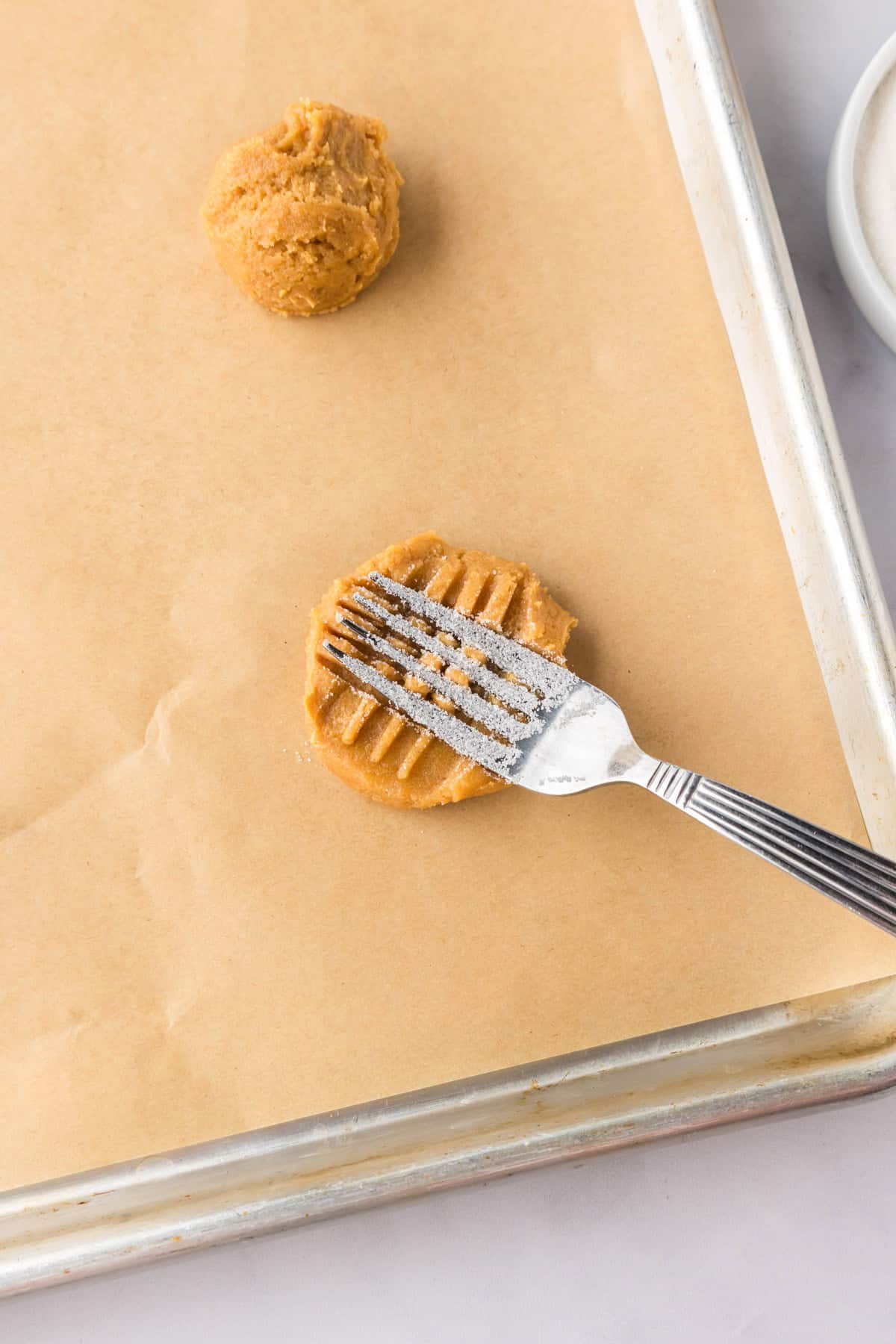A fork presses a 3 ingredient peanut butter cookies dough ball on a parchment-lined baking sheet, creating a crisscross pattern. Another dough ball sits nearby.