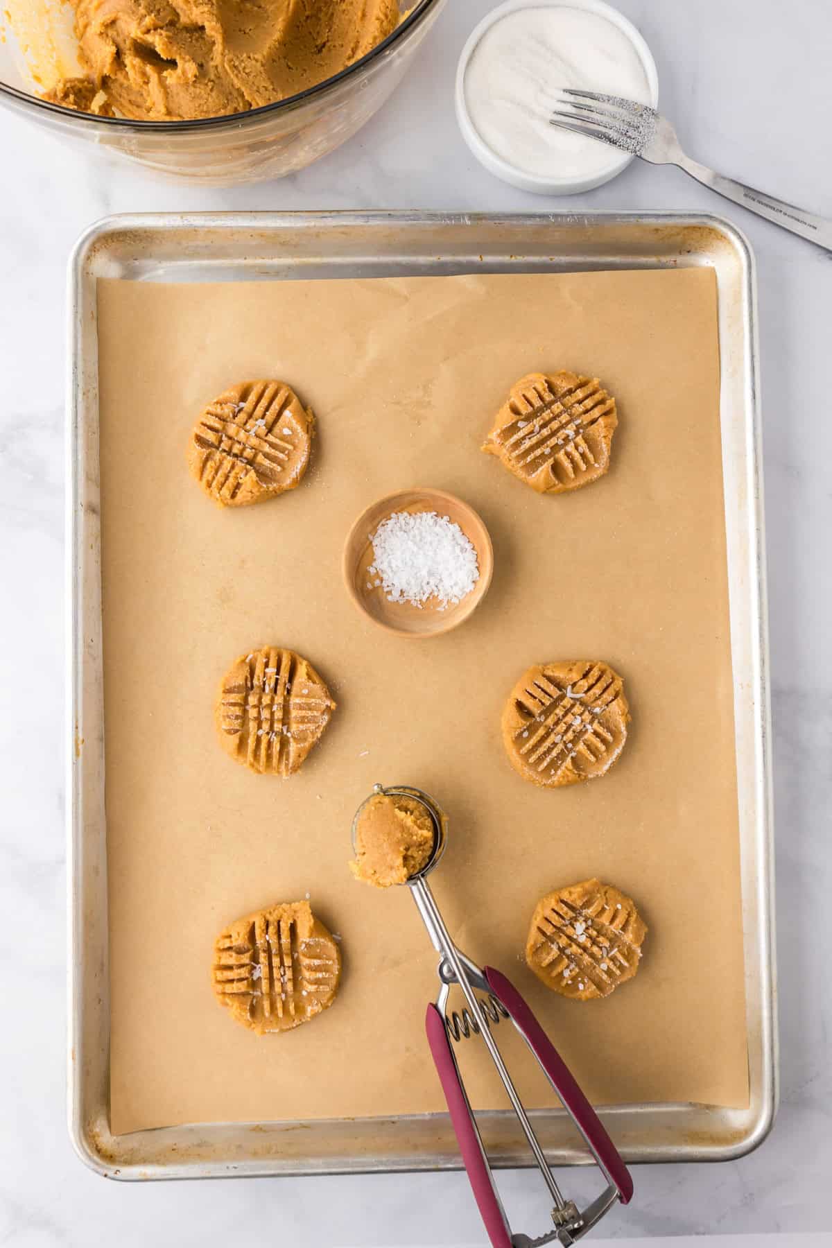 A baking tray lined with parchment paper holds six pressed 3 ingredient peanut butter cookies, a cookie scoop, a small bowl of salt, and some loose salt sprinkled on the cookies.