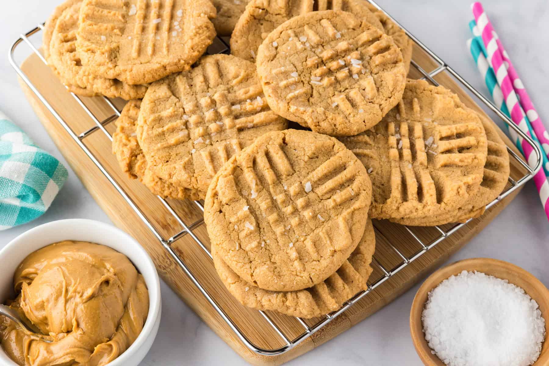A batch of 3 ingredient peanut butter cookies with fork marks rests on a cooling rack, next to a bowl of peanut butter and a small dish of coarse sugar.