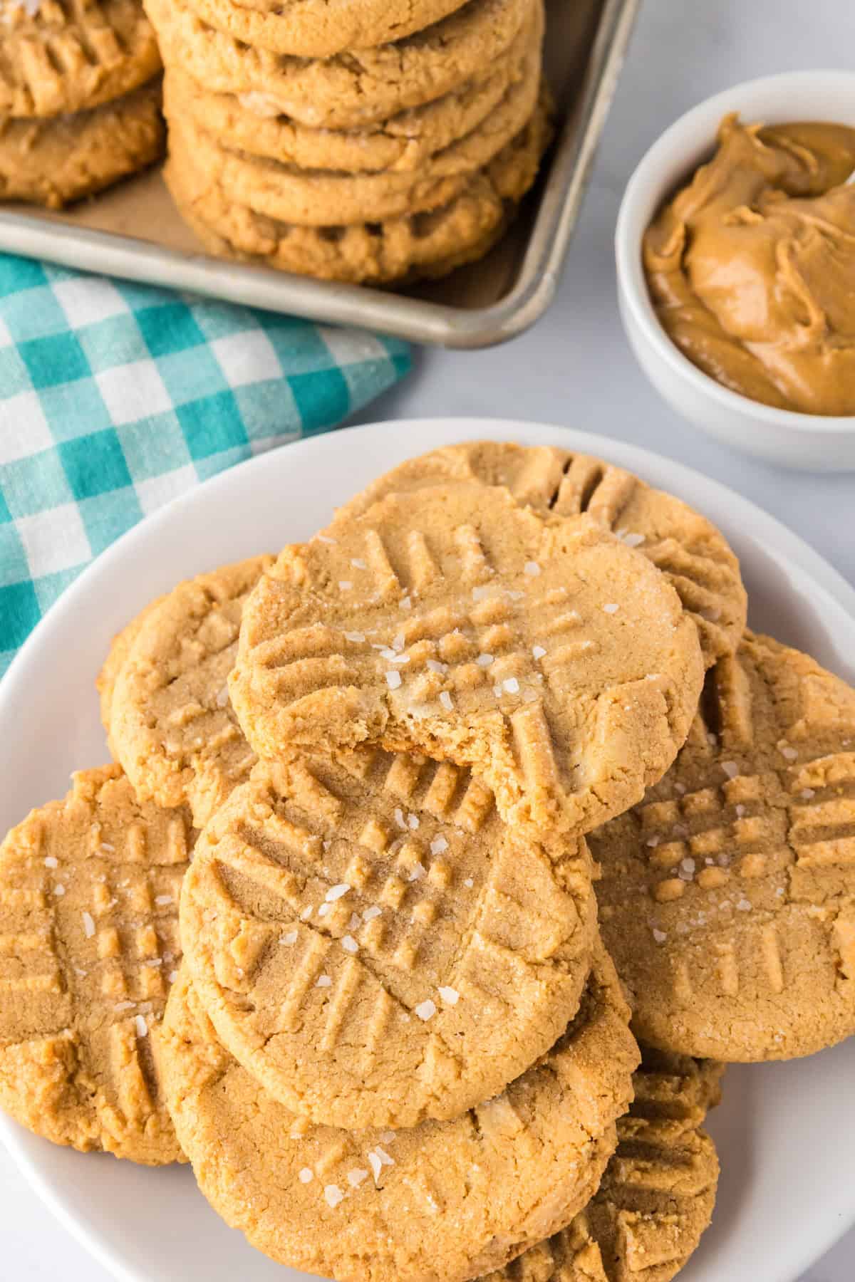 Plate of 3 ingredient peanut butter cookies with a crisscross pattern, sprinkled with coarse sugar, next to a bowl of peanut butter and a stack of cookies on a tray.