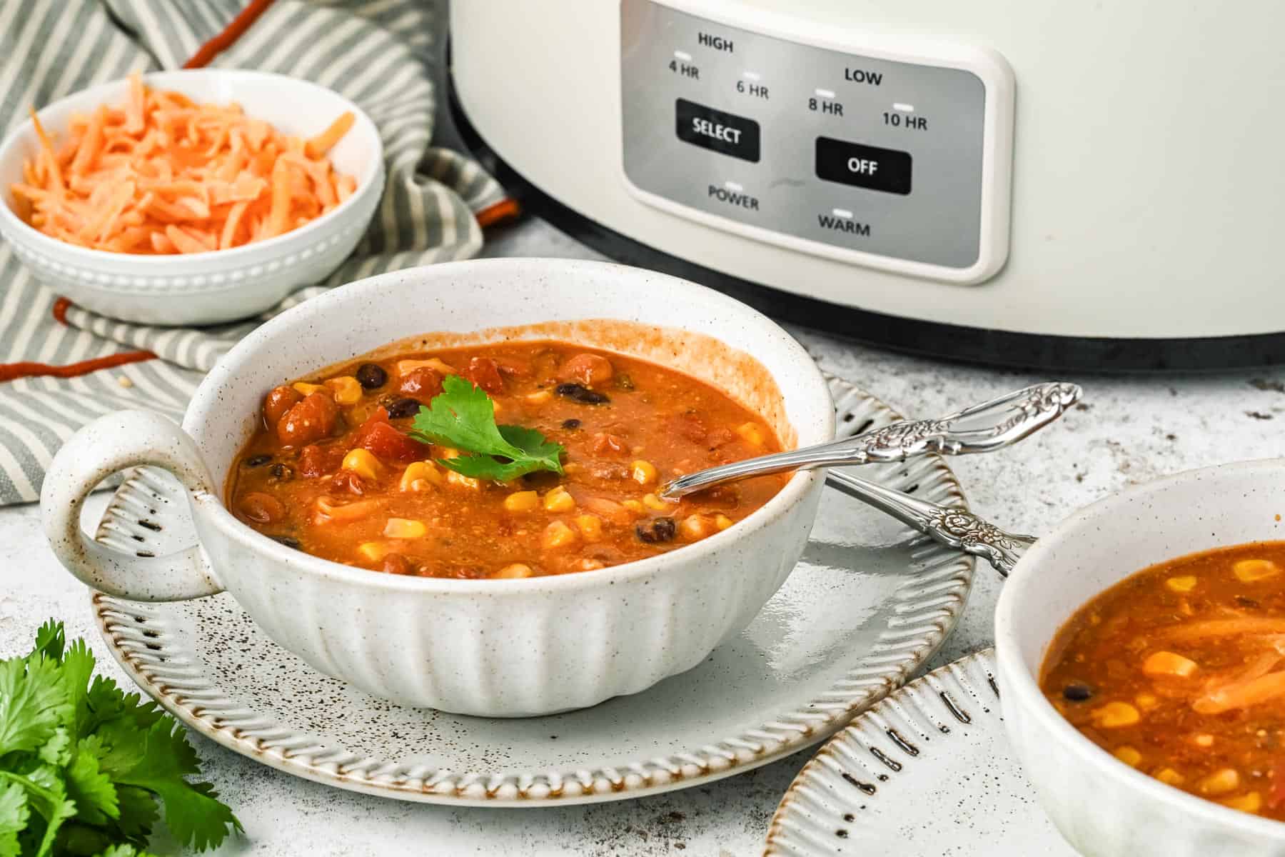 A bowl of crock pot 7 can soup with corn and beans sits on a plate next to a bowl of shredded cheese and a slow cooker, garnished with a sprig of cilantro.