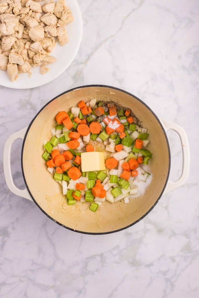 A pot containing chopped carrots, celery, onions, seasonings, and a pat of butter sits on a marble countertop beside a plate of cooked chicken pieces for creamy chicken stew.