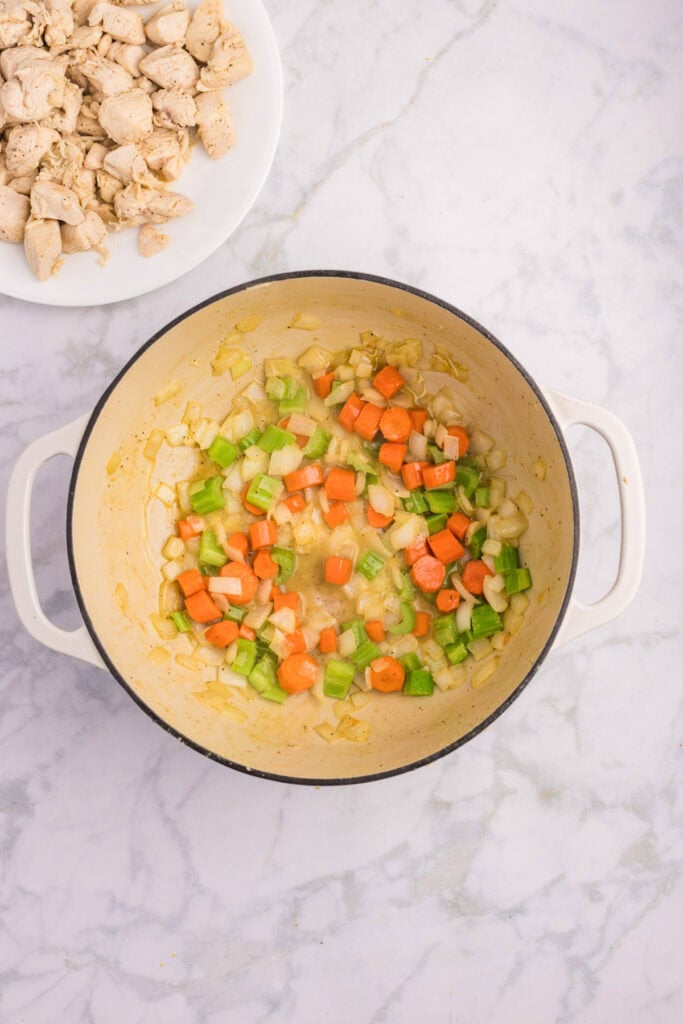 Chopped carrots, celery, and onions are being sautéed in a white pot on a marble countertop; a plate of cooked chicken pieces sits nearby to make a creamy chicken stew.