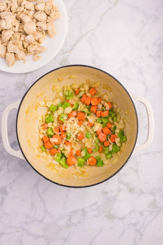 A white pot with chopped onions, carrots, celery, and garlic being cooked with garlic to make a creamy chicken stew.