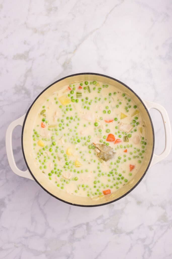 A white pot filled with creamy chicken stew containing peas, diced vegetables, and visible bay leaf/