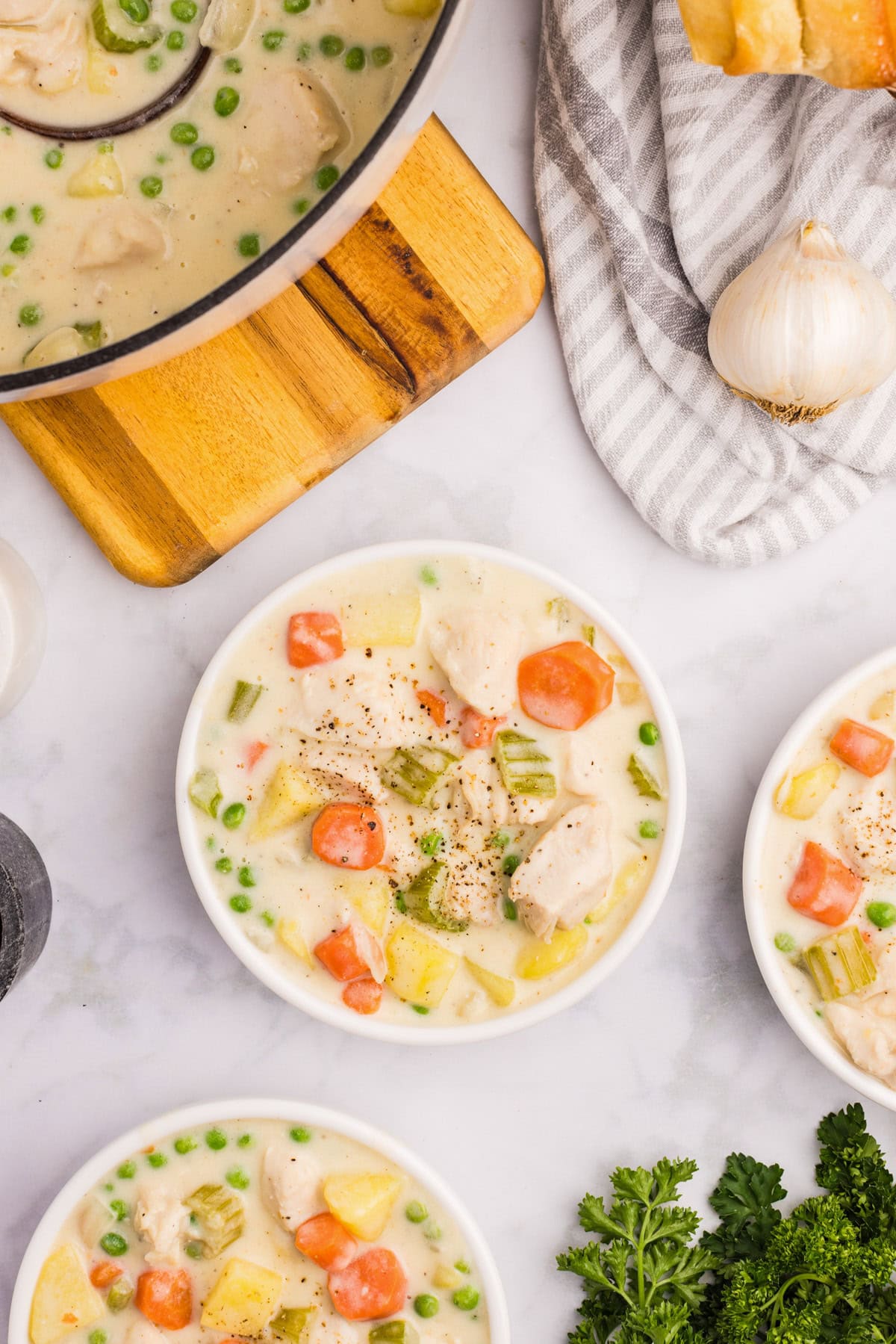 Three bowls of creamy chicken stew with carrots, peas, potatoes, and celery sit on a marble surface, next to a pot of creamy chicken stew, fresh parsley, garlic, and bread.
