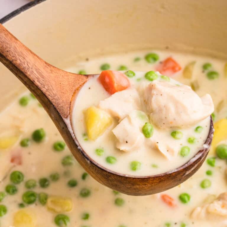 A wooden ladle holding a serving of creamy chicken stew with peas, carrots, and potatoes.