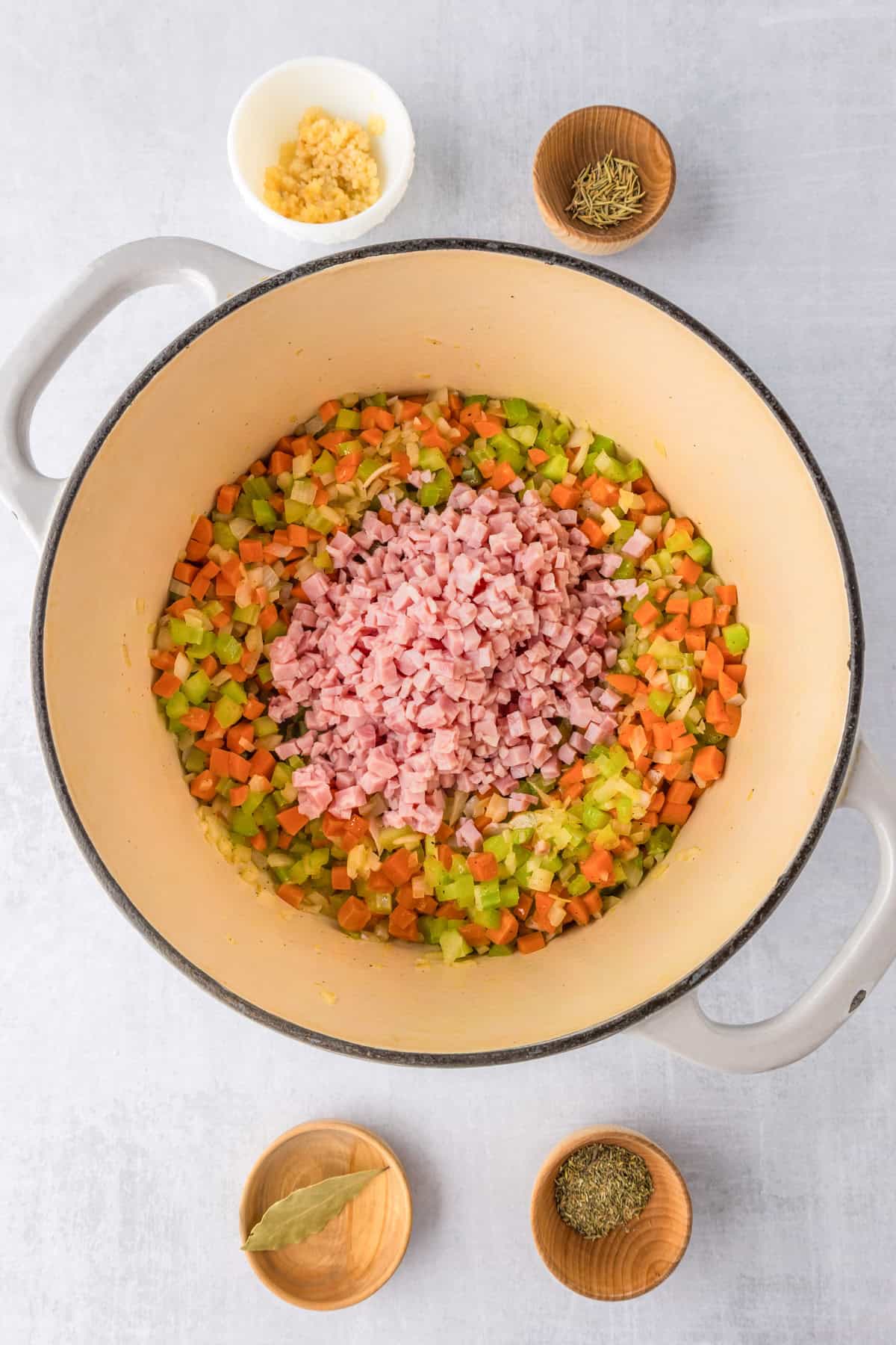 A pot with sautéed diced carrots, celery, onions, and a pile of chopped ham in the center, surrounded by small bowls of minced garlic, bay leaf, and spices, perfect beginnings for a hearty white bean and ham soup.